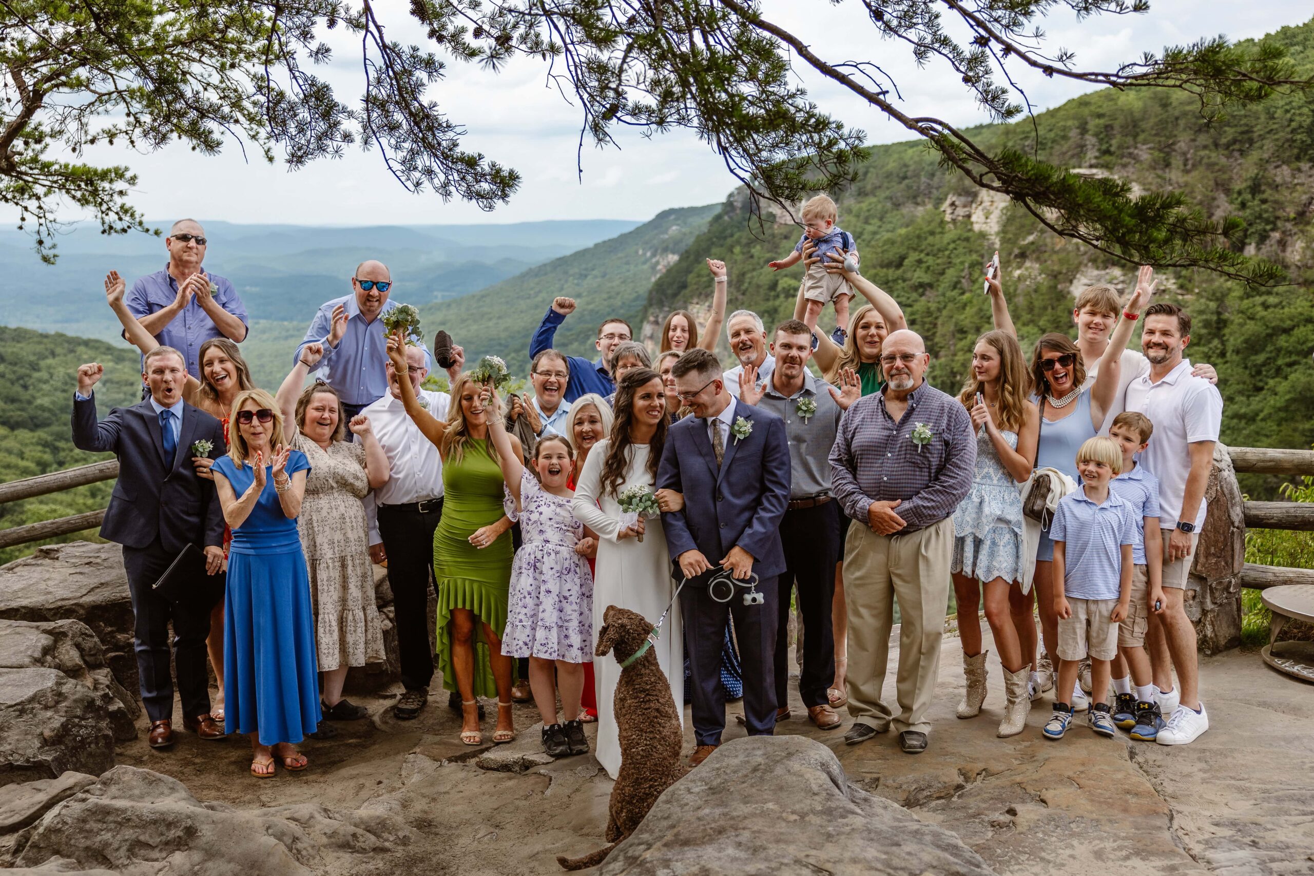 A bride and groom stand in the middle of their loved ones who are cheering for them. They are being cheered on after eloping at Cloudland Canyon. They are standing on an overlook with mountains behind them after eloping with family.