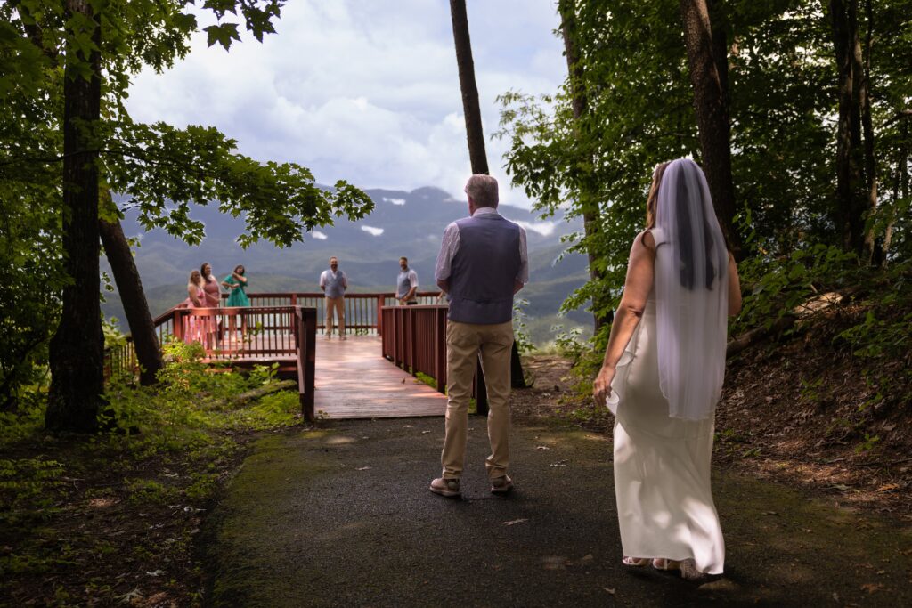 A groom stands with his back facing the camera looking at mountain views. The bride is walking up behind him before eloping in Blue Ridge. In the distance are loved ones waiting to watch them elope.