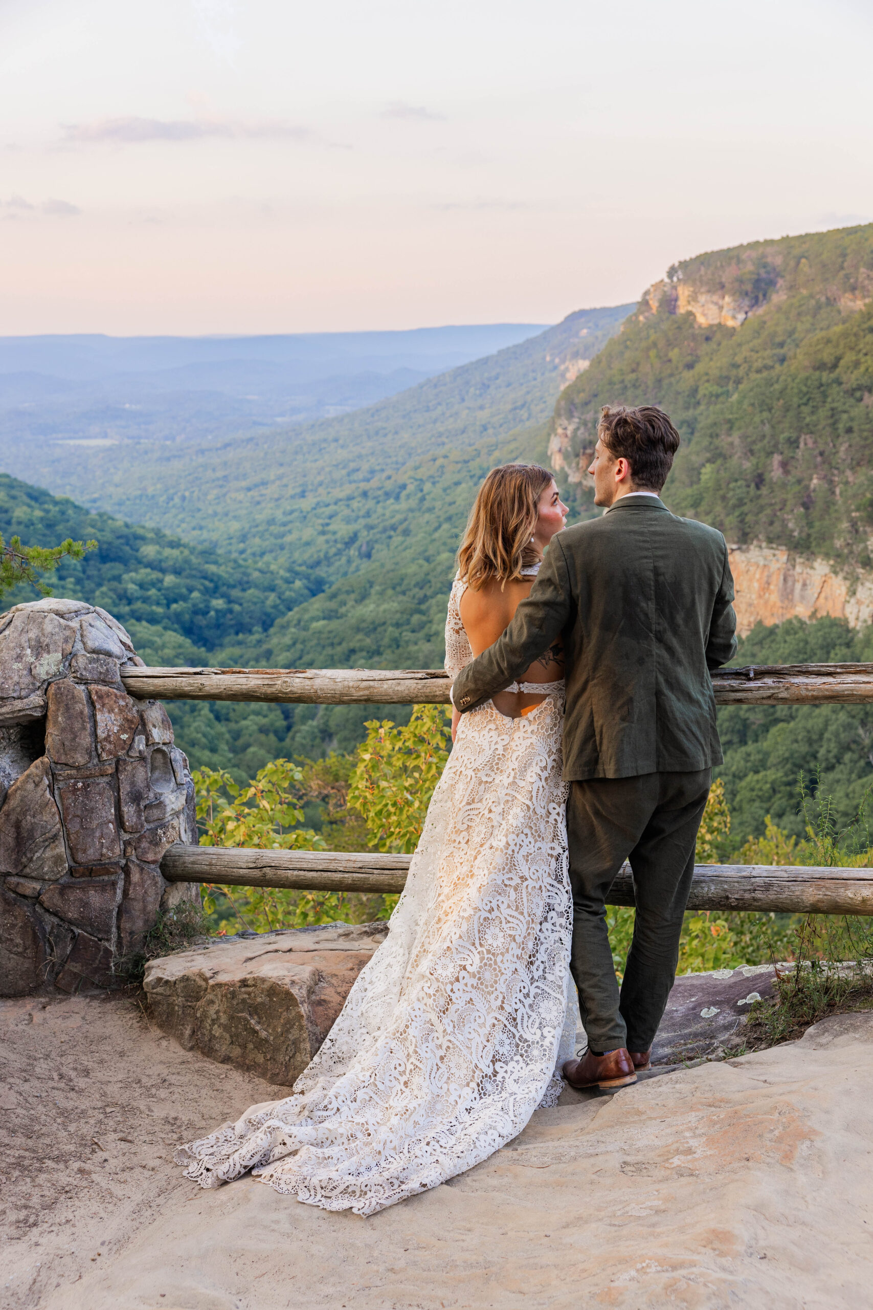 A bride and groom stand with their backs facing the camera looking out at mountain views as the sun sets after their Georgia elopement. The groom has his arm casually around the brides waist as they soak in the views.