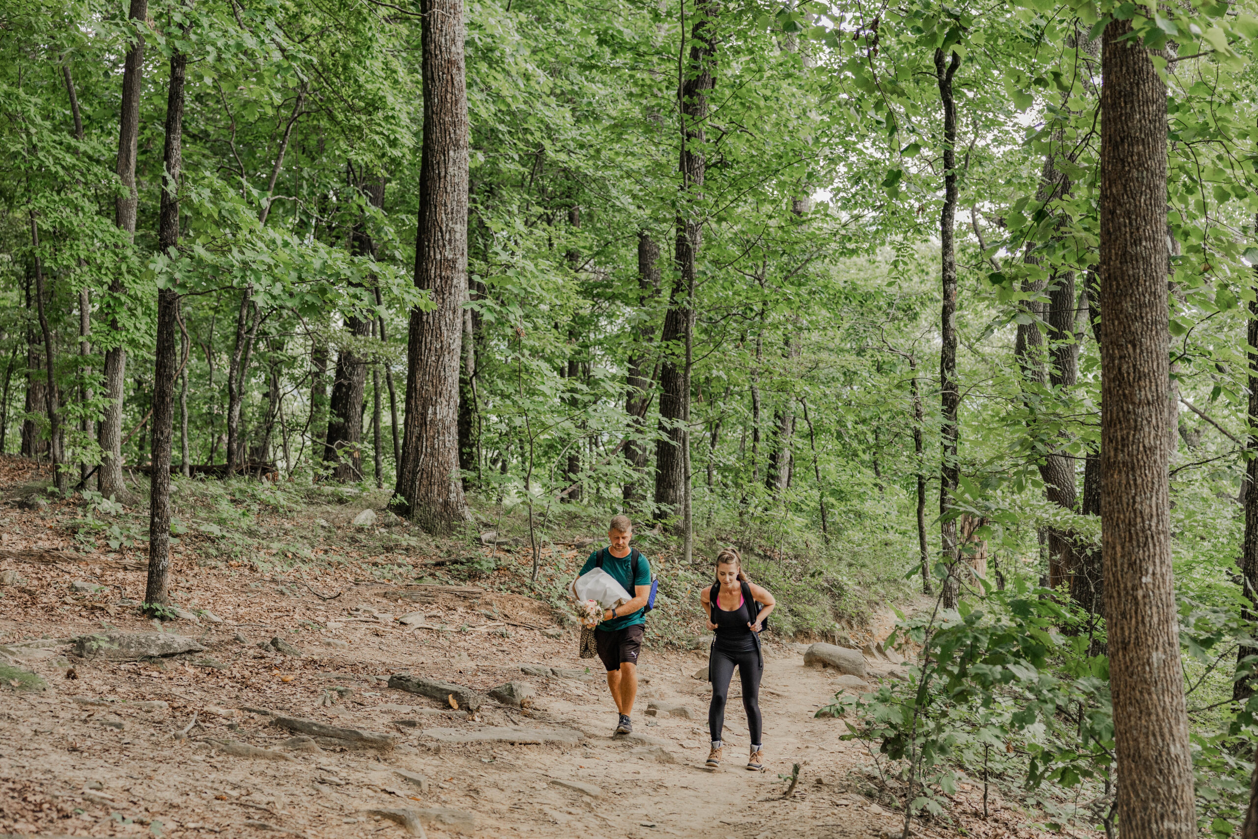 A bride and groom are wearing hiking gear while walking up a trail to their elopement location. The groom is carrying the brides dress. They are on a dirt trail surrounded by vast green pine trees.
