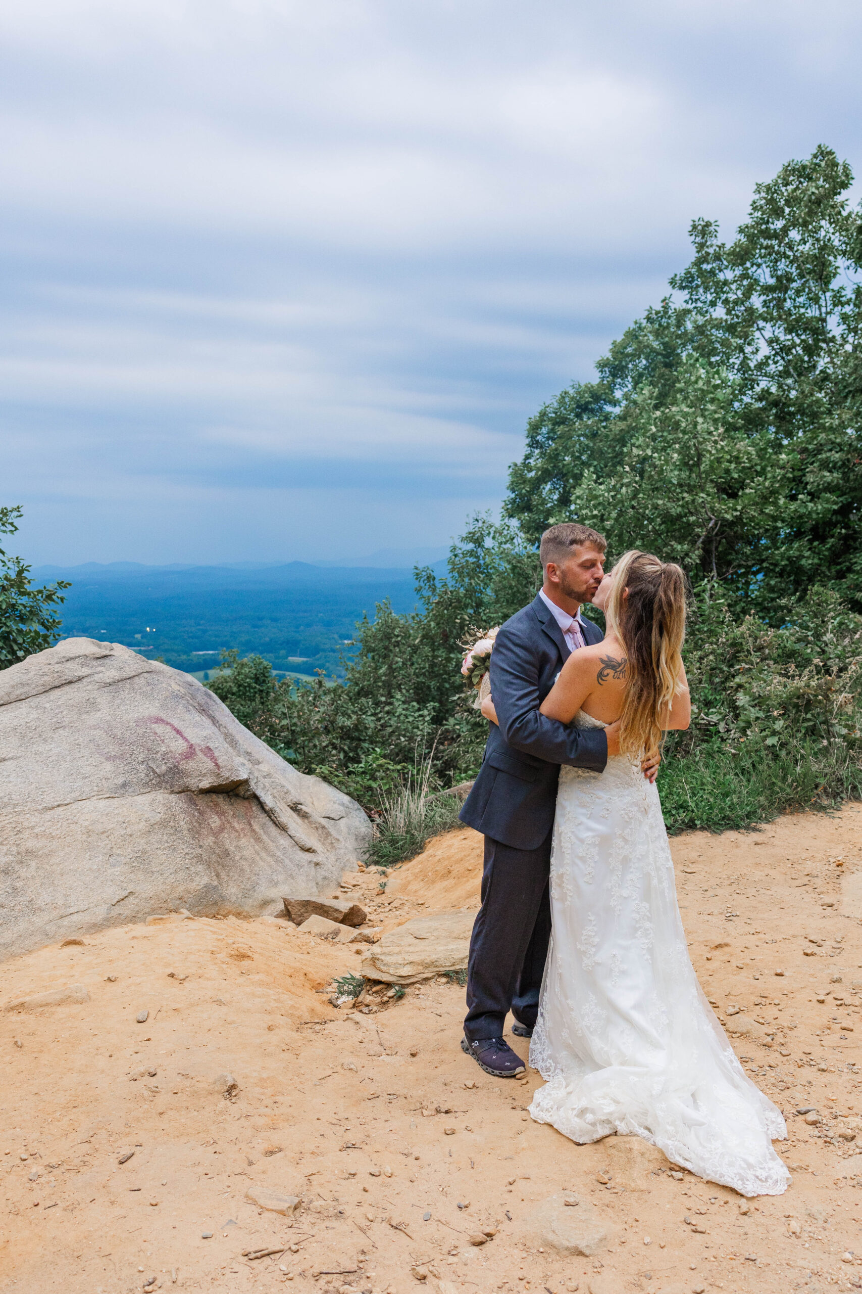 A bride and groom share a kiss after eloping in Georgia. The are holding each other while standing in front of a mountain overlook. The sun has set casting down dark shadows.
