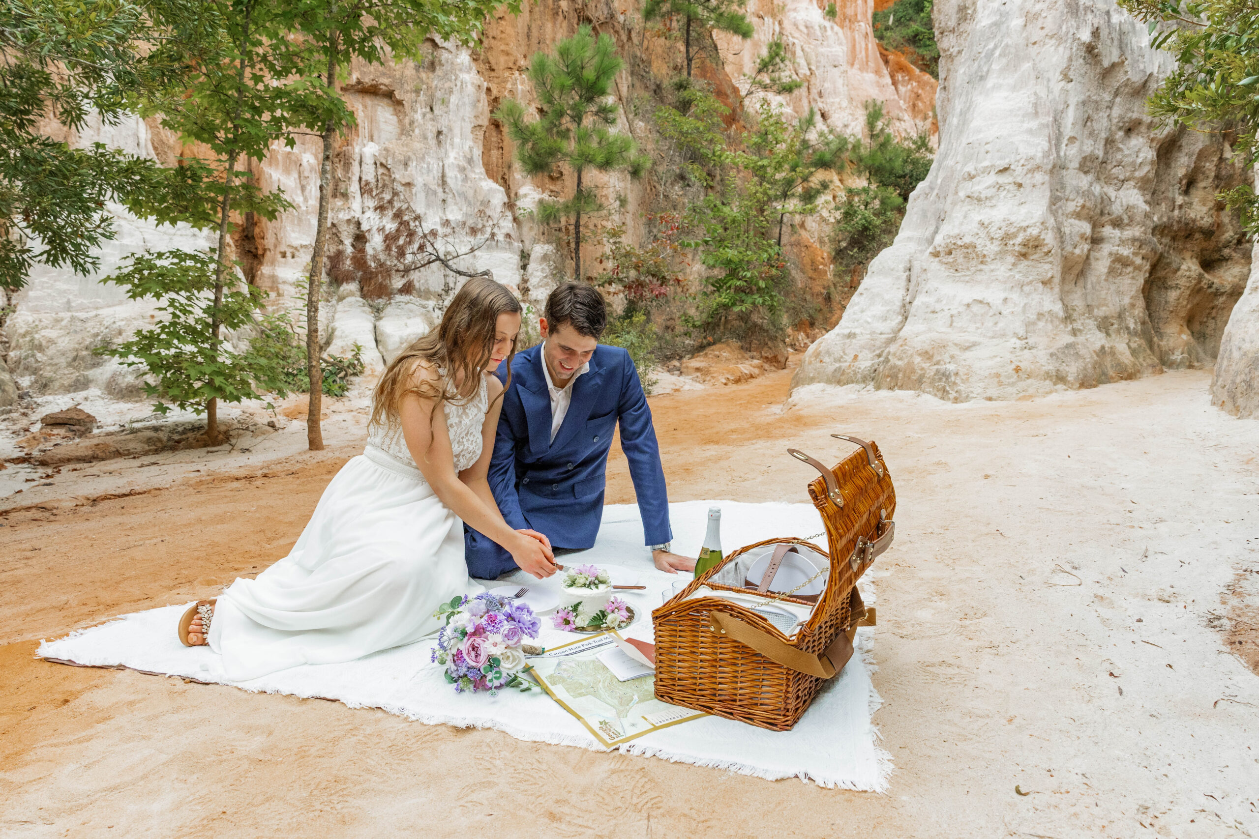 A bride and groom are cutting their cake after eloping in Georgia. They are sitting on a picnic blanket with a basket and bouquet of flowers by them. They are in a red canyon surrounded by red dirt and green trees.