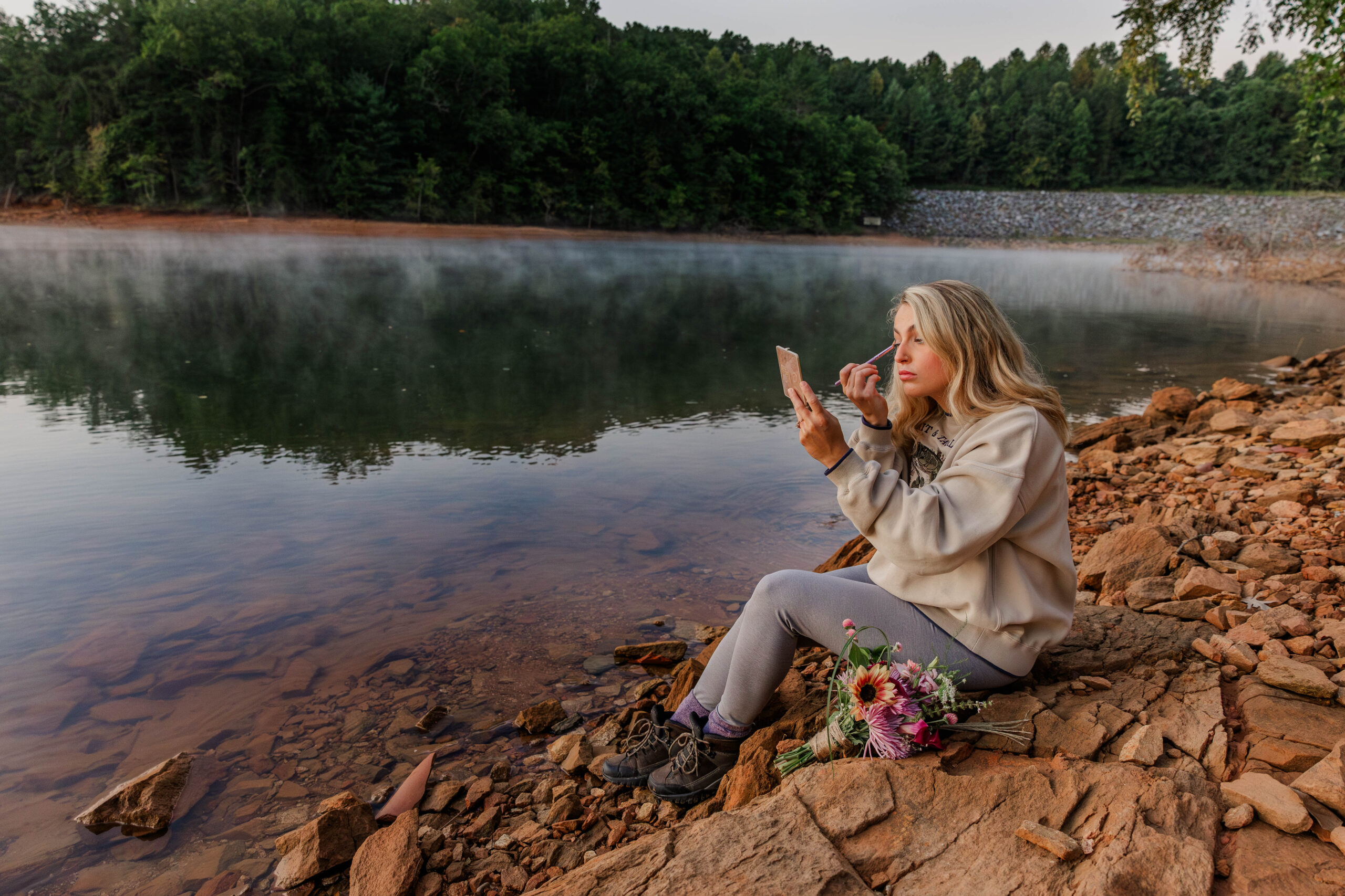 A bride sits on red rocks at the edge of a lake while touching up her makeup. She is using a makeup palette mirror to see herself. She is wearing hiking clothes while her bouquet of wildflowers sits nearby.