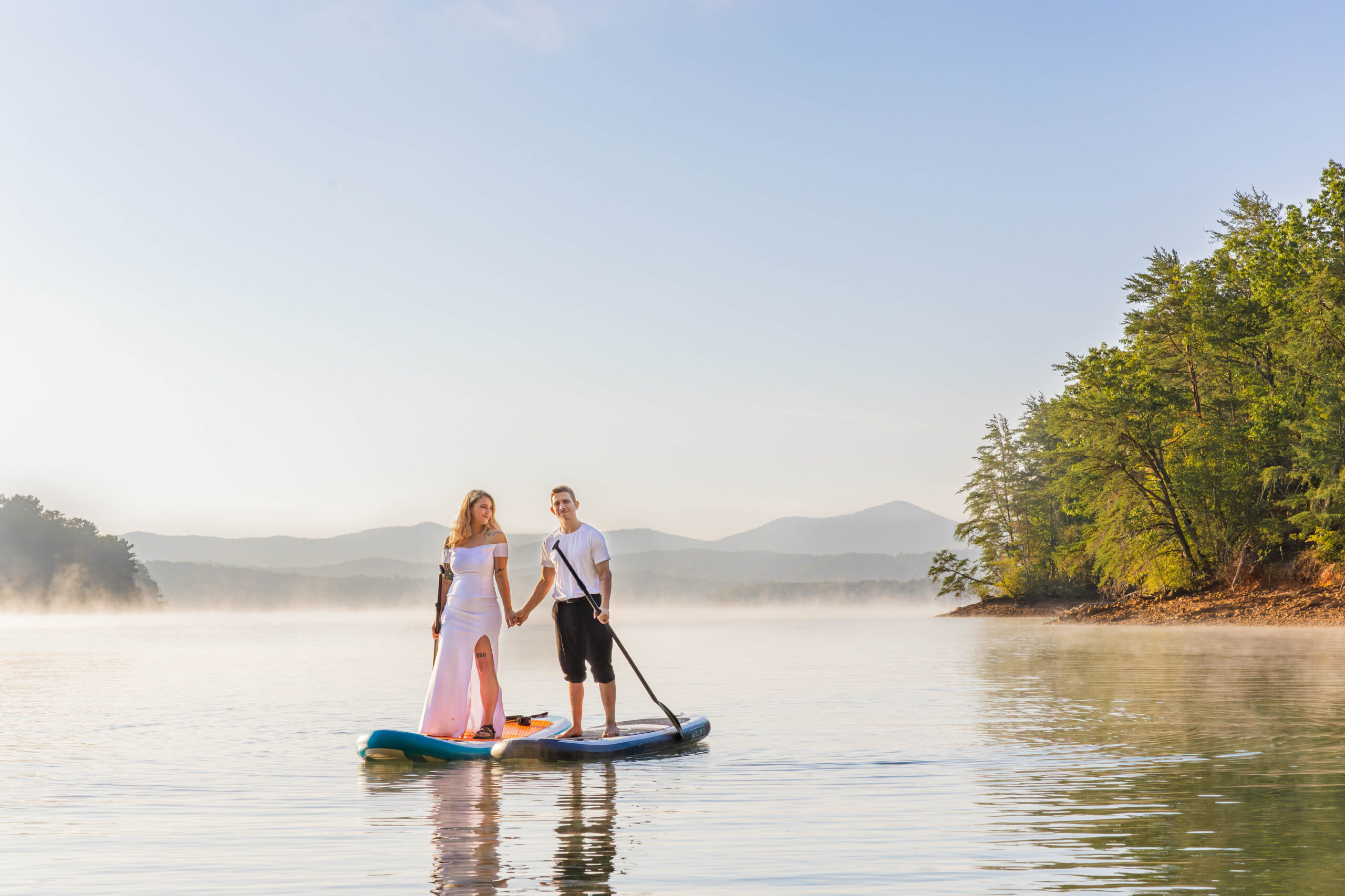 A bride and groom are standing on paddle boards in the middle of a lake holding hands and smiling at the camera. The sun is rising off to the side and there is steam coming off the lake all around them. In the background are vast mountain ranges.