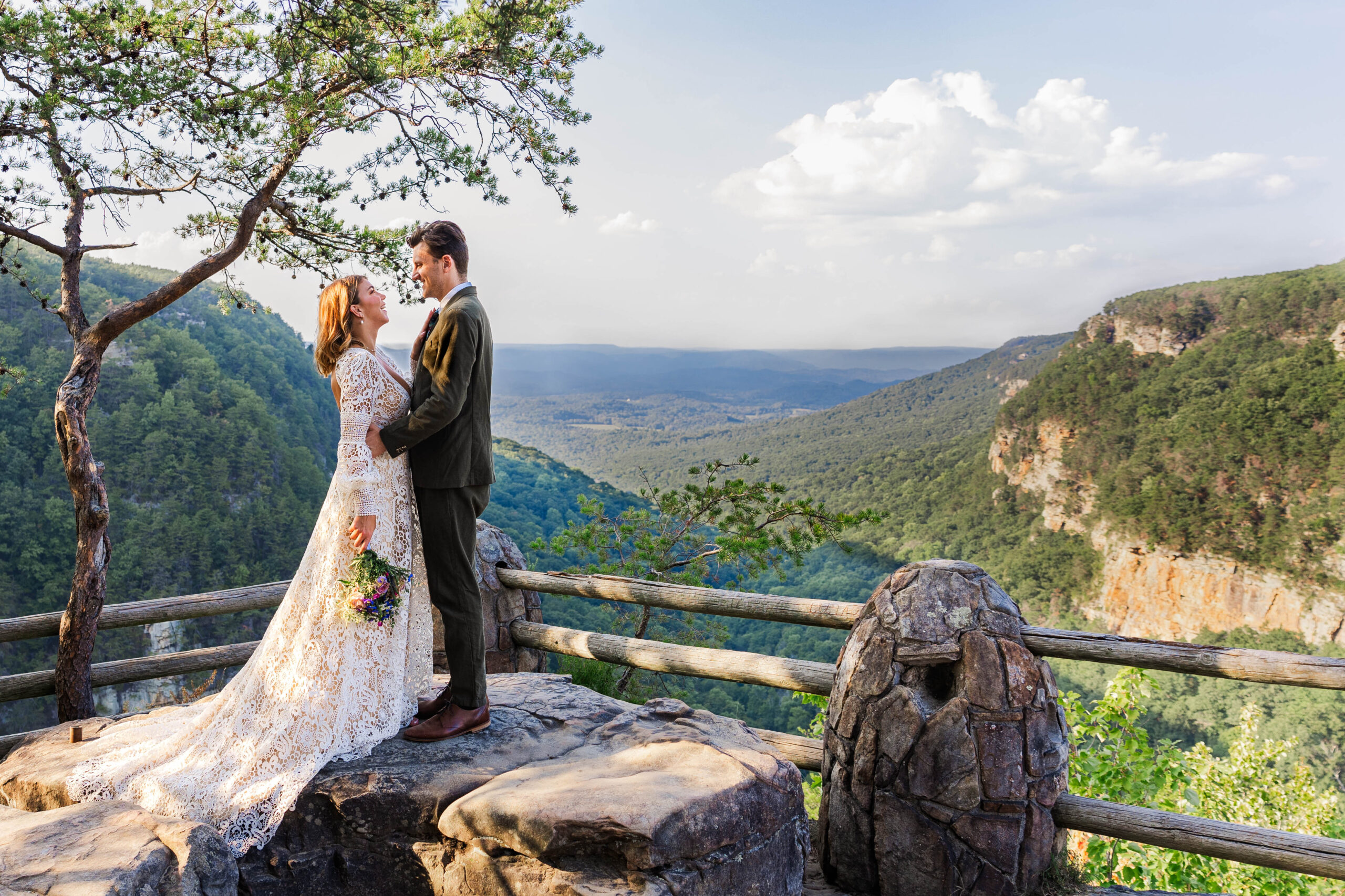 A bride and groom stand looking at each other on top of a rock in front of a mountain overlook. The bride is wearing a long white wedding dress and the groom is in an olive green suit. The background is filled with vast rocky mountains covered in green trees contrasting the blue sky.