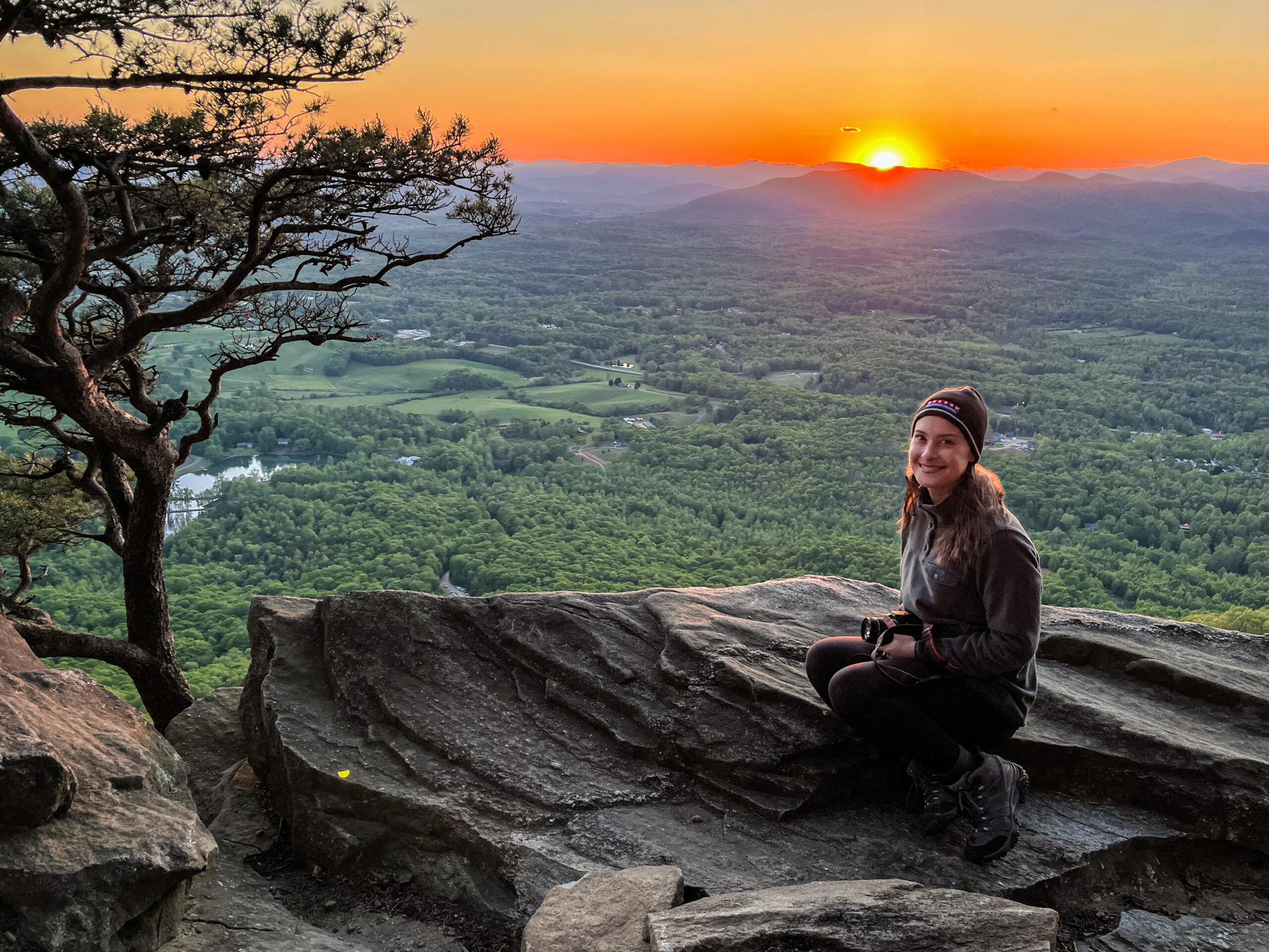An adventure elopement photographer sits on a large rock at the top of a mountain looking out at the landscape while the sun sets in the distance behind her.
