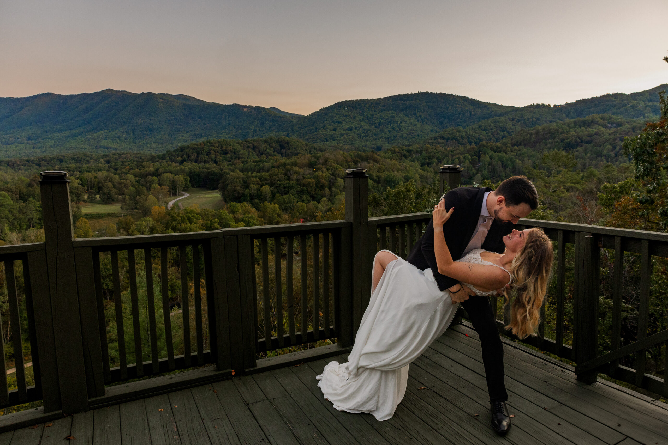 A groom is dipping a bride after eloping in Tennessee. They are standing on a balcony overlooking the smoky mountains as the sun sets