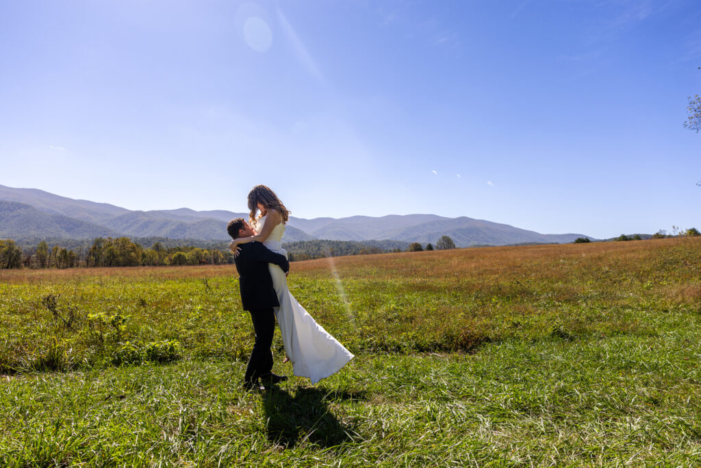 A groom is lifting up the bride as they share a kiss after eloping in the Blue Ridge Mountains. The are standing in a grassy field with a large mountain range behind them