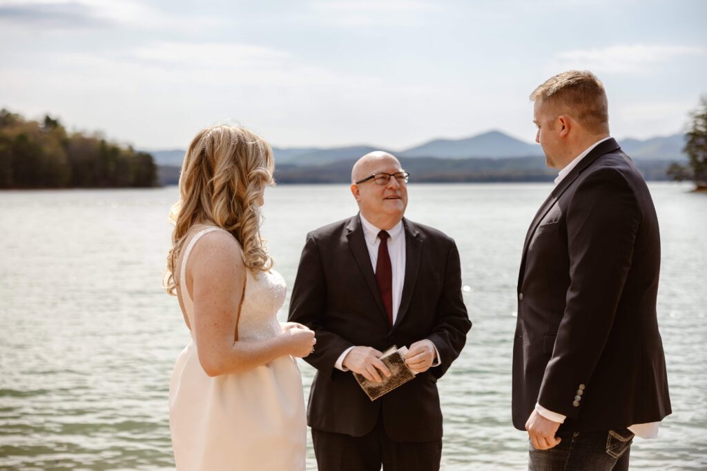 A bride and groom stand with an officiant in front of a lake with a mountain backdrop.
