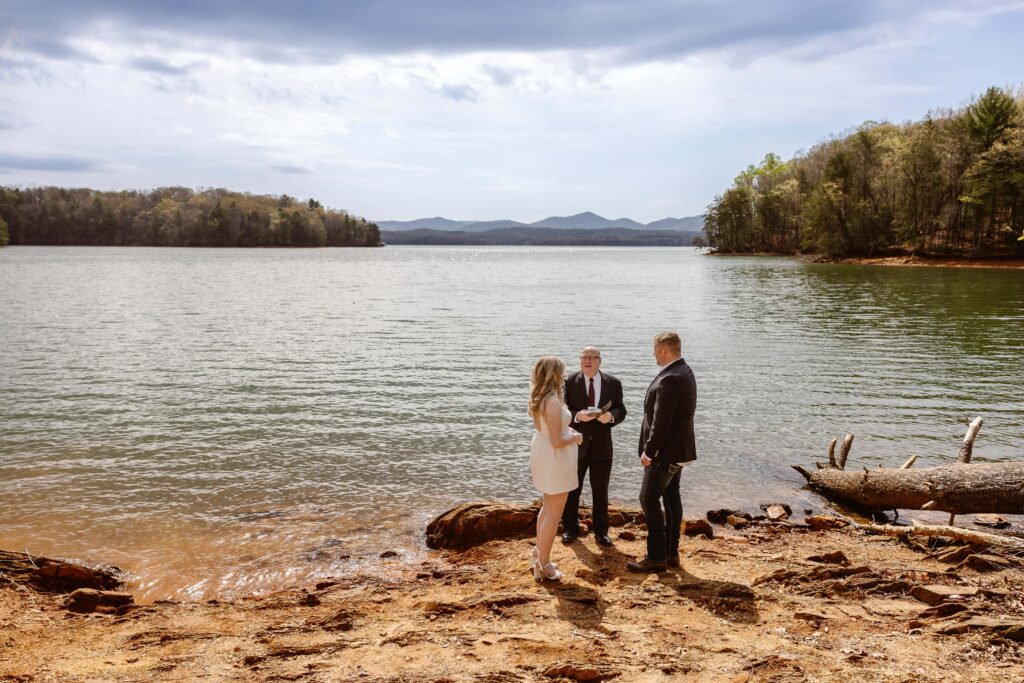 A bride and groom stand on the edge of a lake with mountains and trees in the background. They are standing on a red rocky shore with an officiant behind them during their elopement ceremony. They are eloping in Georgia. The bride is in a short white dress with high heels and the groom is in dark colored jeans and a black suit jacket.
