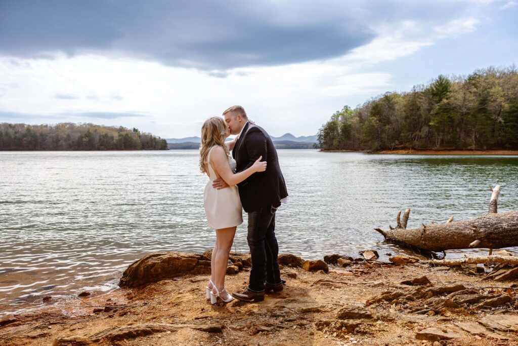 A bride and groom share their first kiss as a married couple after eloping in Georgia. The are standing on a rocky shoreline in front of a lake with mountain views.
