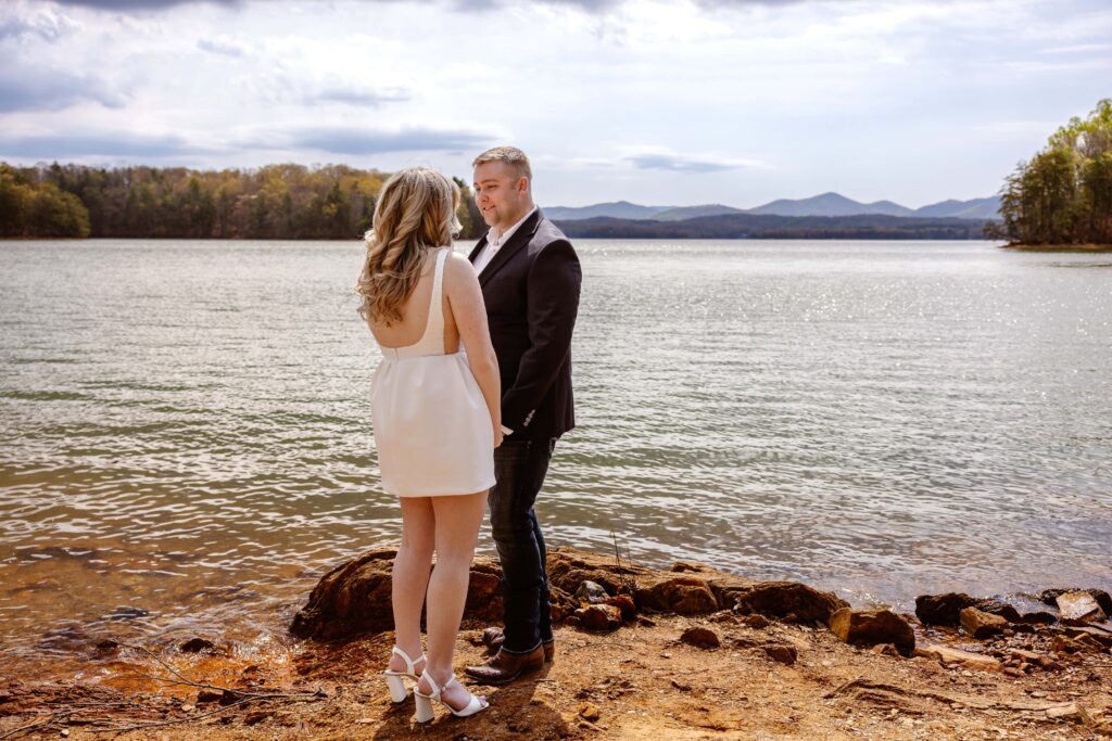 A bride and groom stand on a rocky shoreline in front a vast lake with a mountain backdrop. The brides back is facing the camera as she looks at her new husband. The groom is smiling at his new wife.