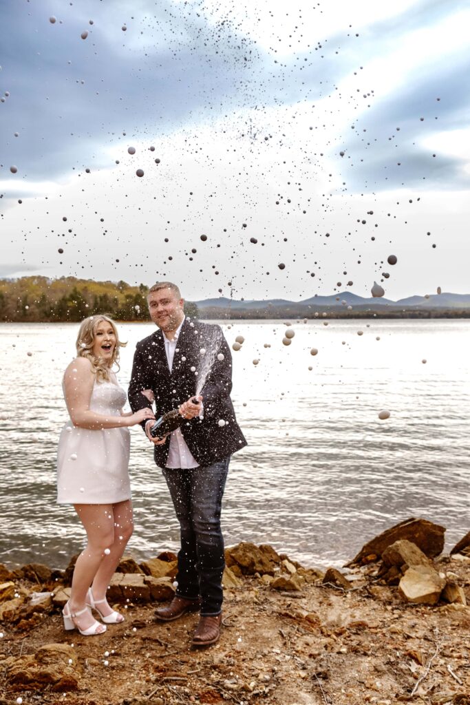 A bride and groom are celebrating their elopement by popping champagne on the edge of a lake with mountain views. The groom is smiling as he sprays the champagne and the bride is smiling and laughing.