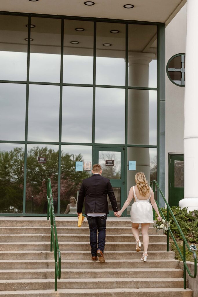 A bride and groom walk up the steps of the county courthouse while holding hands to return their signed marriage license after their elopement ceremony.