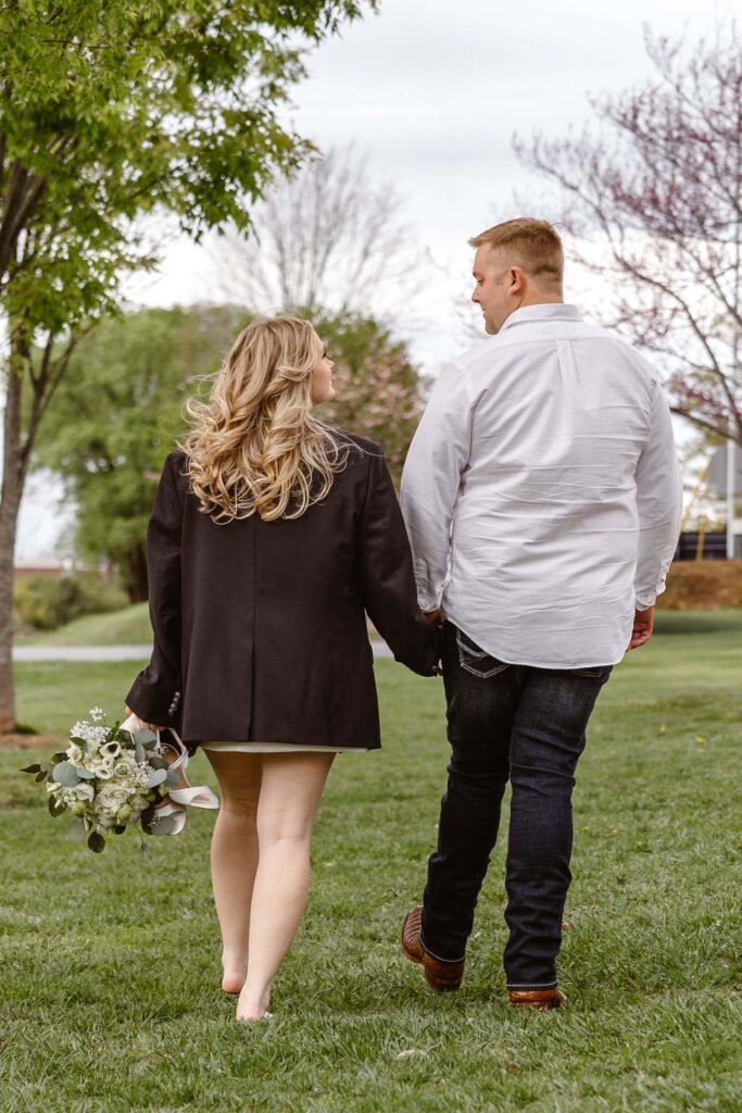 A bride and groom are looking at each other lovingly after eloping. The are holding hands walking away from the camera through grass.