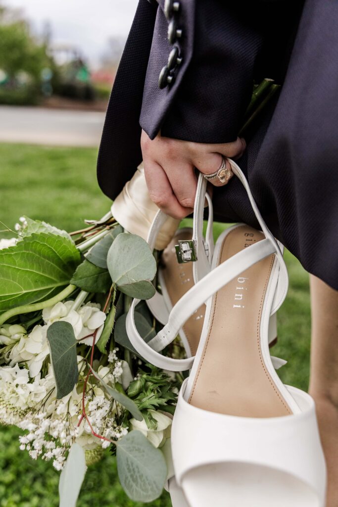 A bride is wearing her grooms suit jacket while walking through grass carrying her heels and bouquet of white wedding flowers.