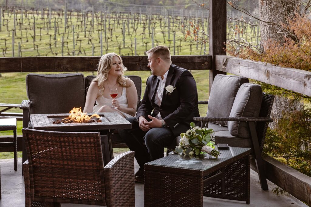 A bride and groom sit by a fire in front of a vineyard of grapes enjoying wine after their elopement. They are sharing an intimate moment with the bride smiling at her new husband.