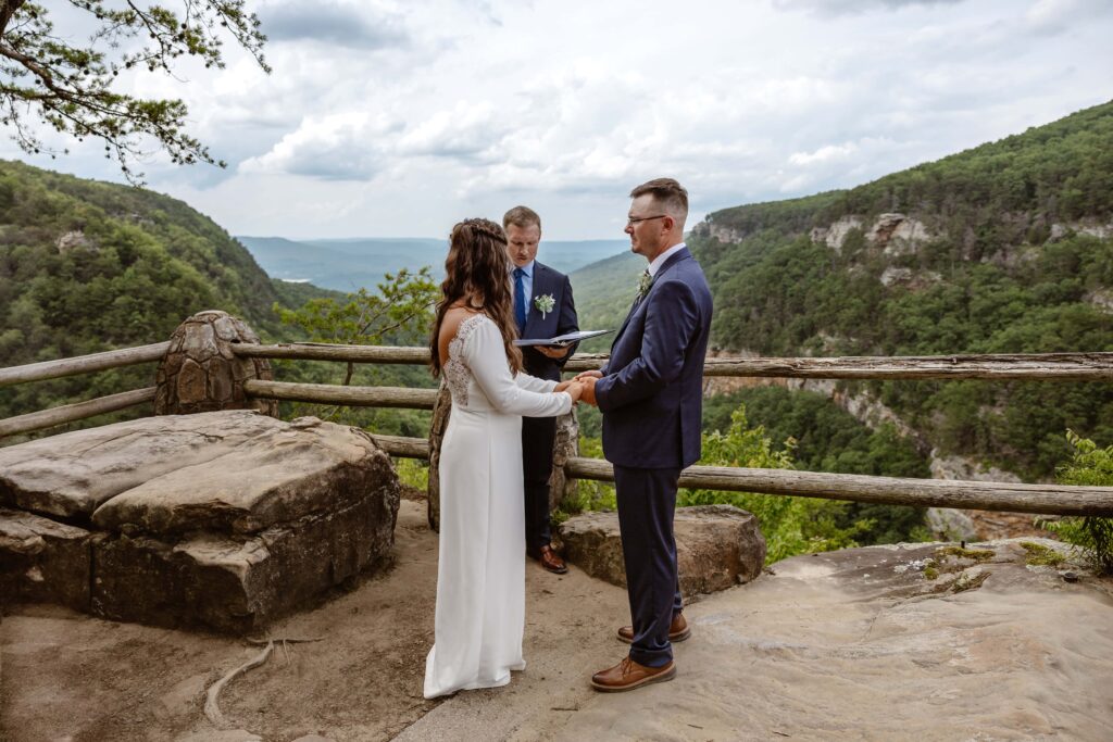 A bride and groom are standing on the edge of a mountain overlooking a vast green mountain range during their elopement ceremony.