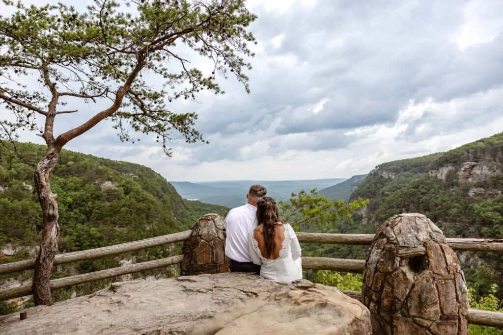 A bride and groom sit side by side with their backs facing the camera looking out at a mountain view after eloping. The brides head is gently resting on the groom shoulder.