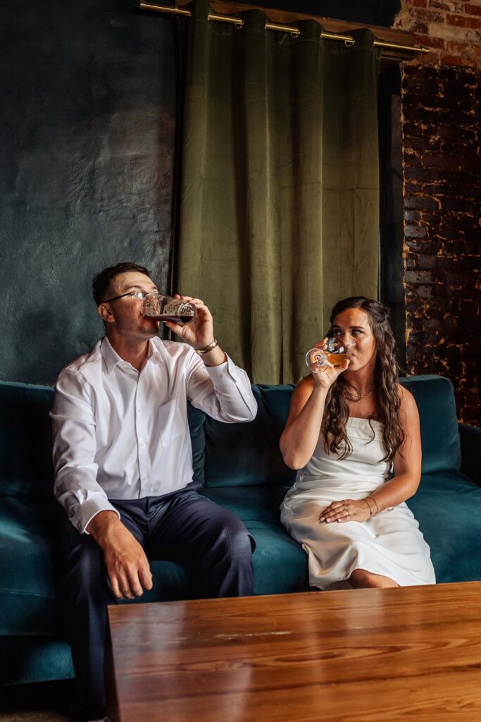 A bride and groom sit side by side drinking beer from Oddstory brewing on their elopement day.