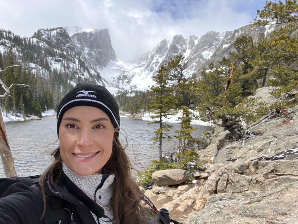 A woman is smiling at the camera taking a selfie. she is standing in front of a lake with a vast snow covered mountain range in the background.