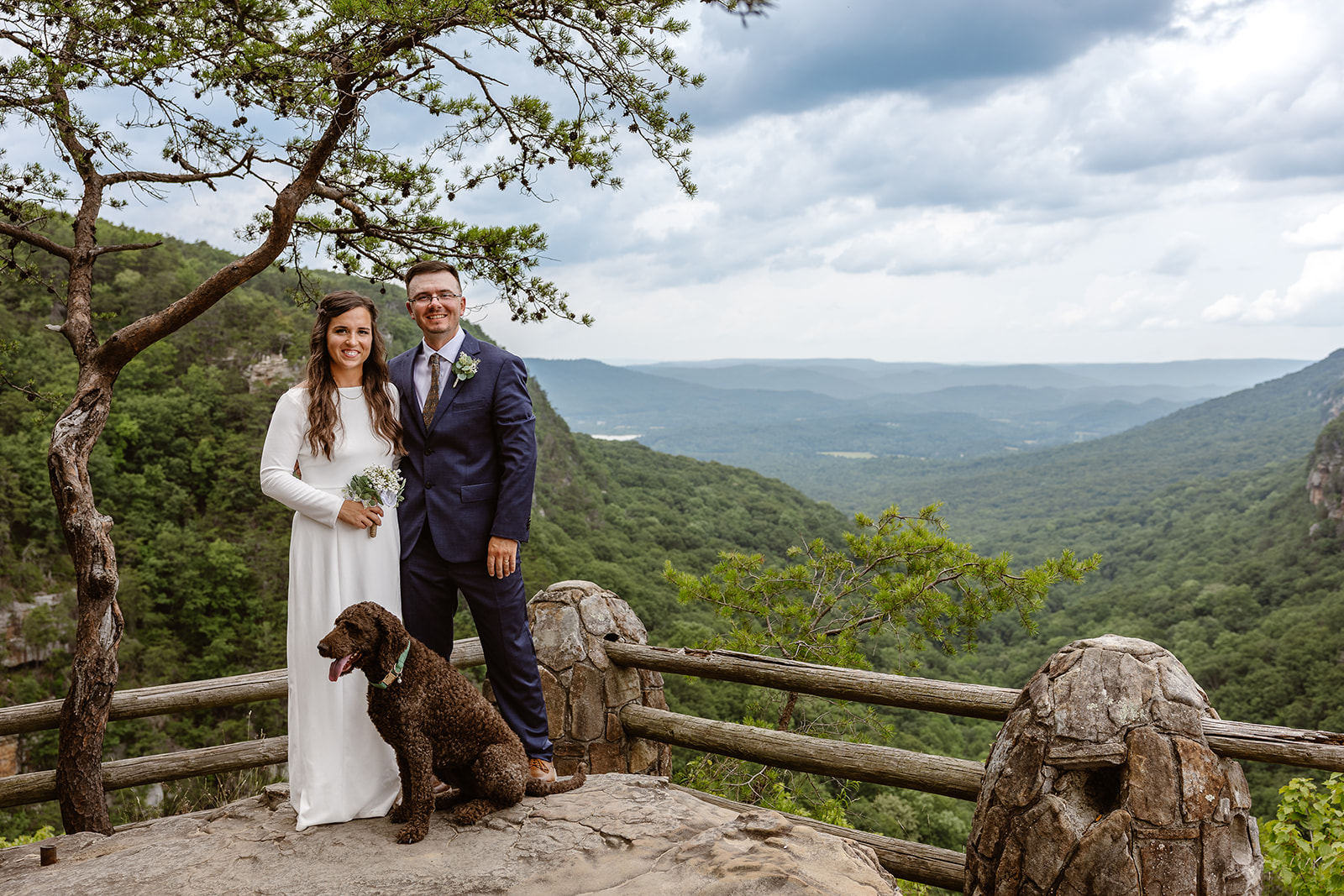 bride and groom take a photo with their dog at the edge of the mountains
