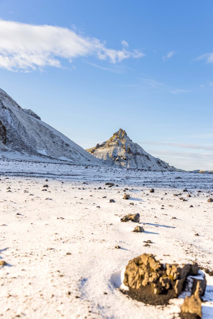 A snow covered ground in Iceland is covered in rocks leads to a large and vast pointed mountain in the distance.