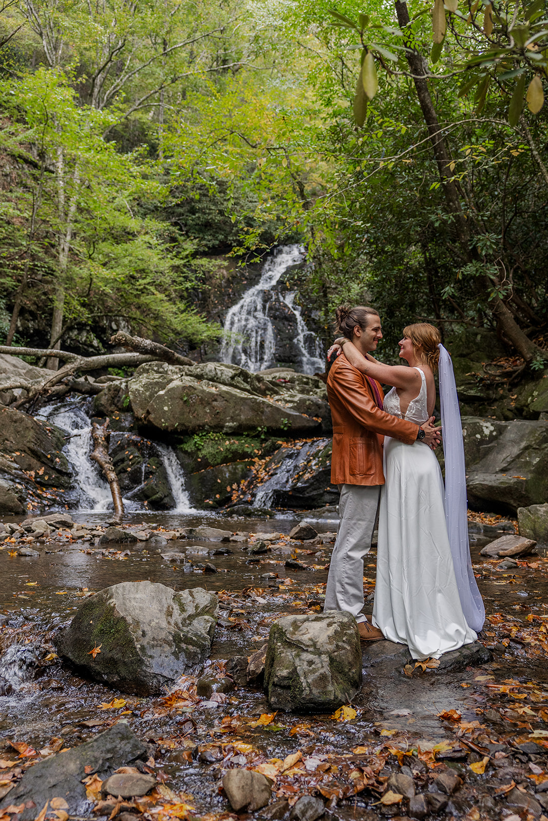 A bride and groom share an embrace looking lovingly at each other. They are getting ready to elope in front of waterfalls in western north carolina