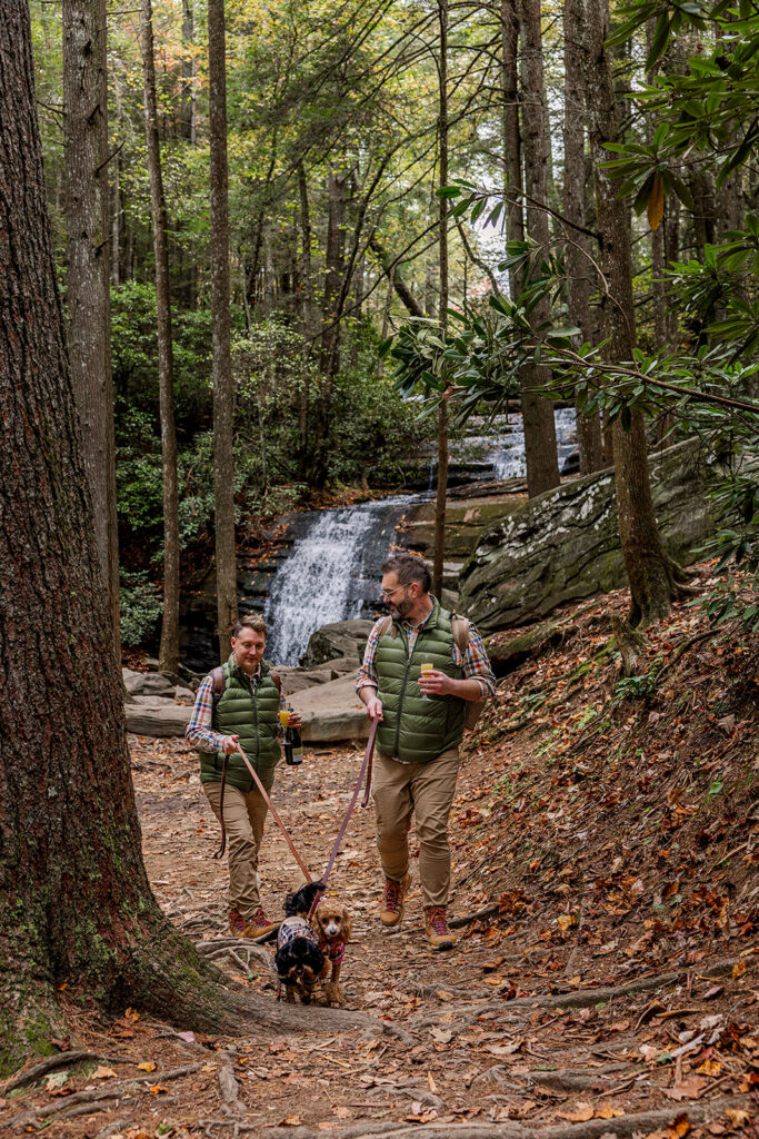 Two grooms are walking their dogs through the woods leaving a waterfall after exchanging private vows and having a picnic. They are having the elopement day of their dreams after spending months elopement planning