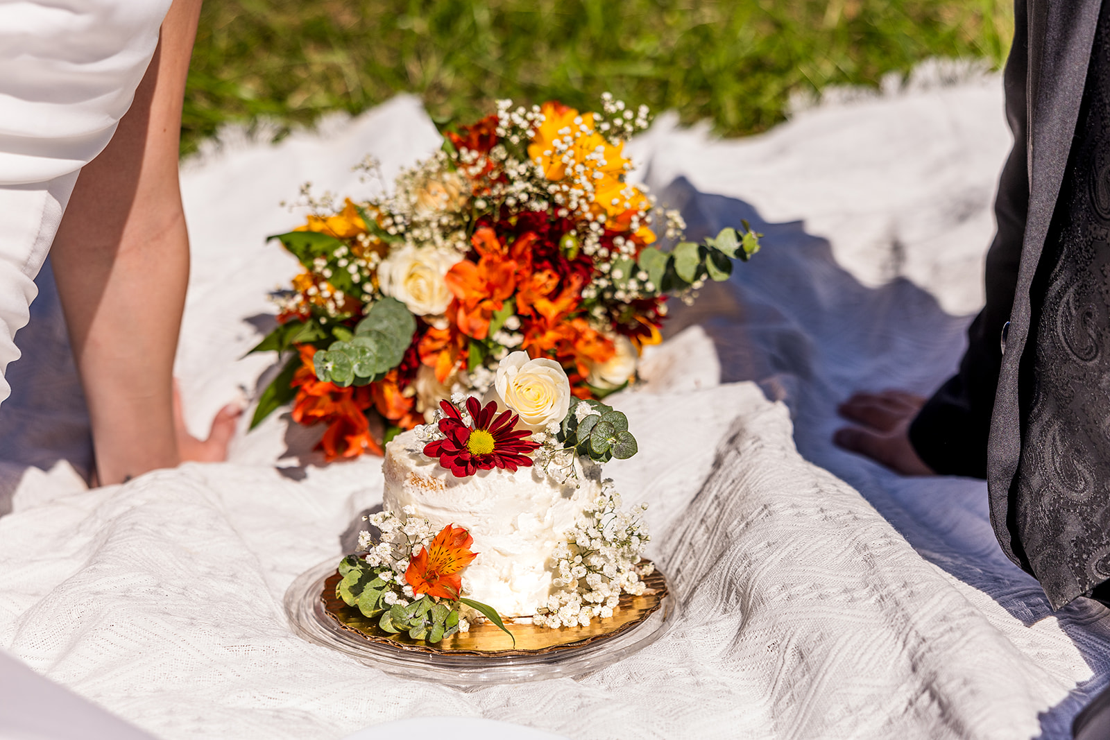 stunning wedding cake sits on the picnic blanket with the bride and groom