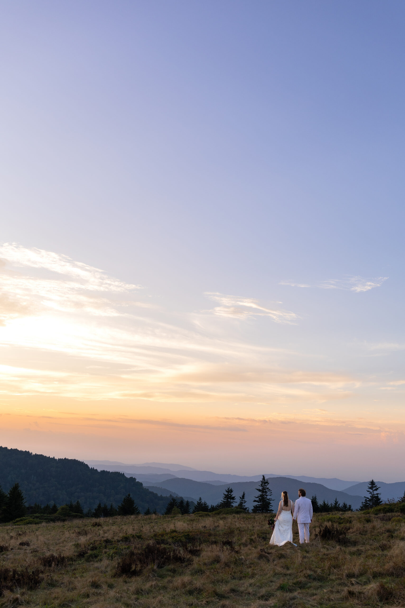 A bride and groom stand on a grassy mountain top walking off into the pastel sunset holding hands with mountain ranges in the distance after their Roan Mountain Elopement