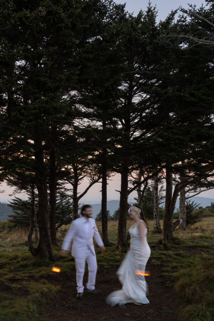 A bride and groom dance in the trees as the sun goes down holding lanterns leading to blurry movement photos. They are celebrating getting married.