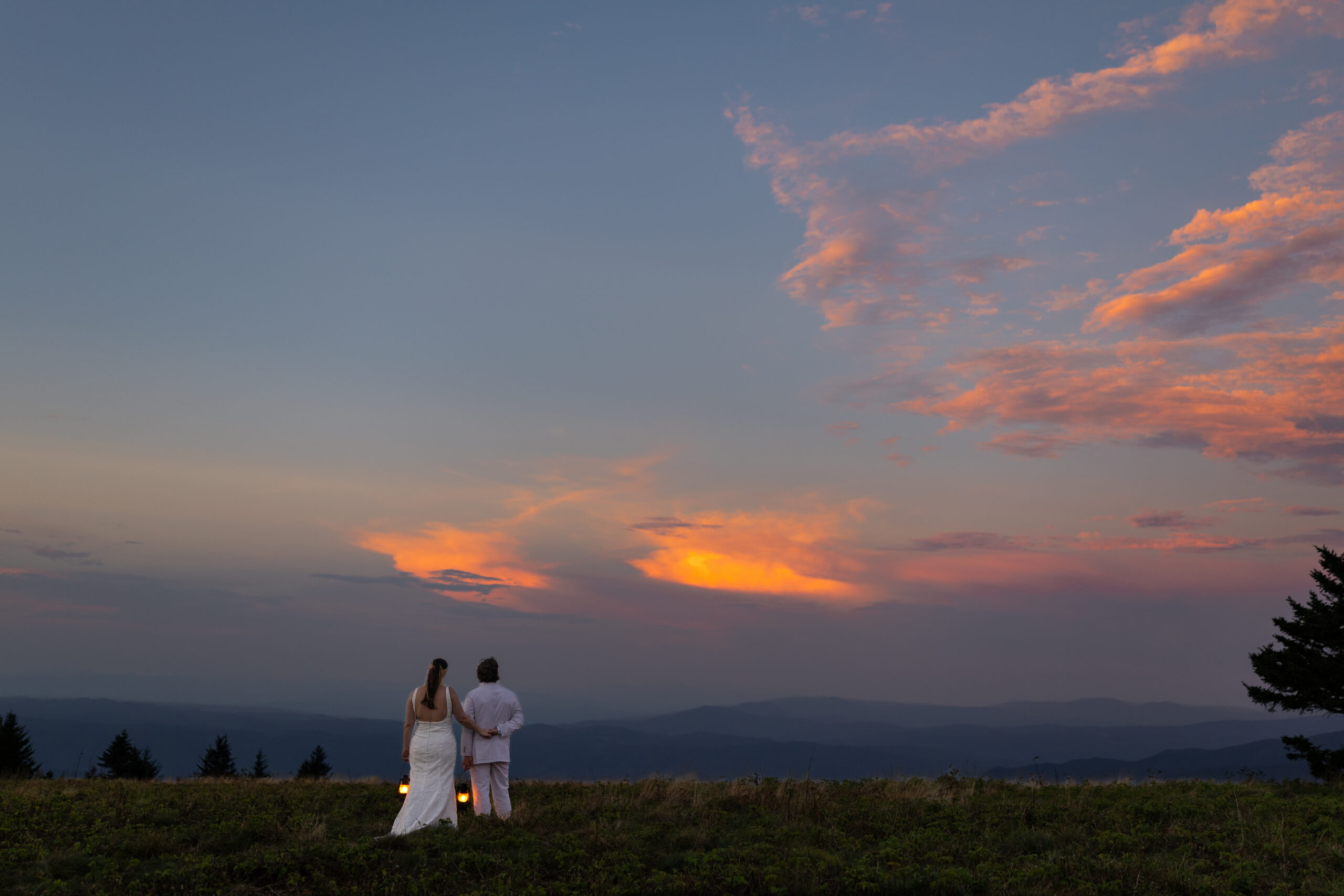 A bride and groom stand with their backs facing the camera holding lanterns and looking out as the sunset turns the sky bright pink and orange over a vast blue mountain range after eloping in Roan Mountain