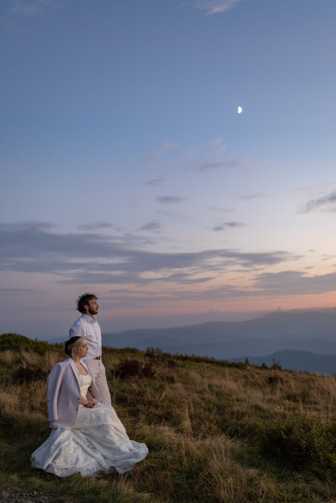 A bride sits in her wedding dress on a rock draped in her grooms suit jacket as he stands beside her. They are looking out at the sunset. In the background you can see the moon rising and pastel pink and blue colors setting over the vast mountain range.