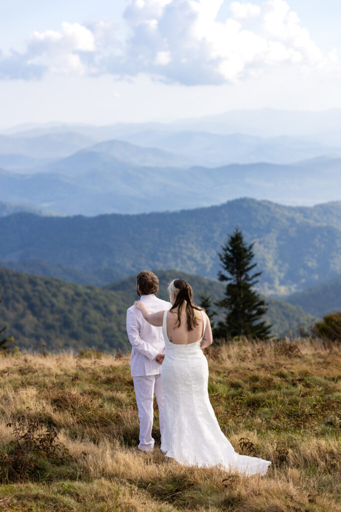A groom is looking out at mountains as the bride stands behind him tapping him on the shoulder. He is about to turn around and see his bride for the first time on their Roan Mountain elopement day.