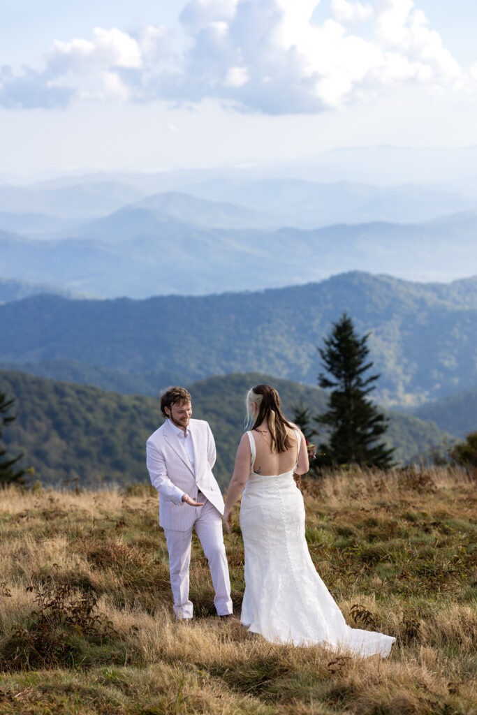 A groom is seeing his bride dressed up in her wedding dress for the very first time. They stand in a grassy field on the edge of a mountain top with 360-degree mountain views. He is smiling at her and gesturing to her wedding dress.