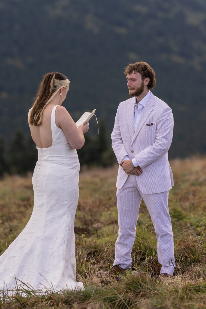 A bride is reading her wedding vows to the groom. The brides back is facing the camera and you can see the handwriting in her vow book. The groom is looking at her holding in a sob as the bride reads the emotional vows.