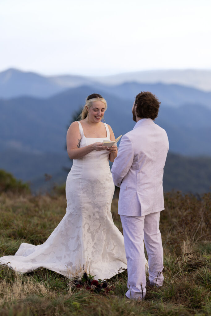 A bride is smiling as she reads her vows to her groom as they stand in a grassy field atop Roan Mountain with mountains in the background.